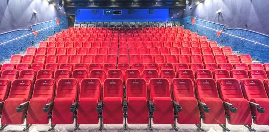 Ein leeres Kino mit Reihen roter Sitze vor einer großen Leinwand, blauen Wänden und beleuchteten Ganglichtern.