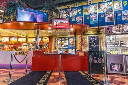 Kassenschalter in der farbenfrohen Lobby eines Kinos mit sichtbaren Filmplakaten, Kiosken, elektronischen Anzeigen und Absperrseil.