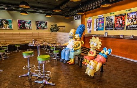 Statuen der Simpsons-Figuren stehen auf einer Bank in einem farbenfrohen Café im Retro-Stil mit Filmplakaten an den Wänden und grün gepolsterten Hockern um kleine Tische.