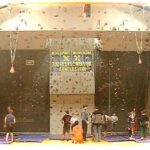 Kinder und Erwachsene stehen und bereiten sich auf das Klettern in einer Kletterhalle mit Kletterwänden und Sicherheitsmatten vor.