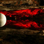 Eine große weiße Kugel schwebt vor einer felsigen, höhlenartigen Oberfläche, die mit rotem Licht beleuchtet ist und Reflexionen nach unten wirft.