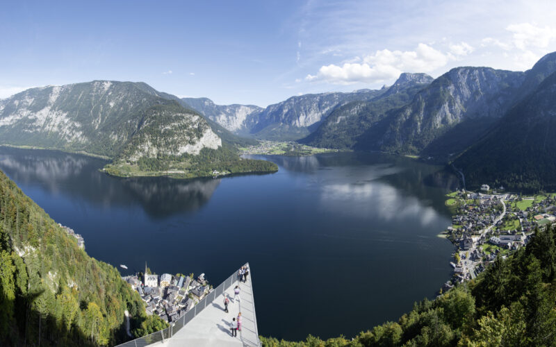 Panoramablick auf einen von Bergen umgebenen See mit einer Aussichtsplattform im Vordergrund und einer kleinen Stadt am Ufer.