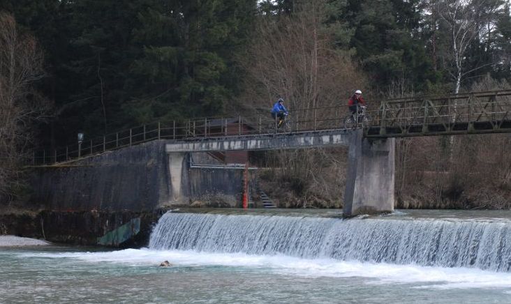 Zwei Radfahrer überqueren eine schmale Brücke über einen Fluss mit einem kleinen Wasserfall. Unten im Wasser nahe der Wasserfallkante ist eine Person zu sehen.
