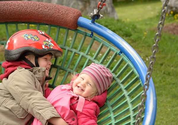 Zwei kleine Kinder, eines davon mit Helm, liegen lächelnd zusammen auf einer großen runden Schaukel auf einem Spielplatz.