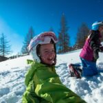 Zwei Kinder mit Helmen und Winterkleidung spielen an einem sonnigen Tag im Schnee, umgeben von Bäumen und Bergen.