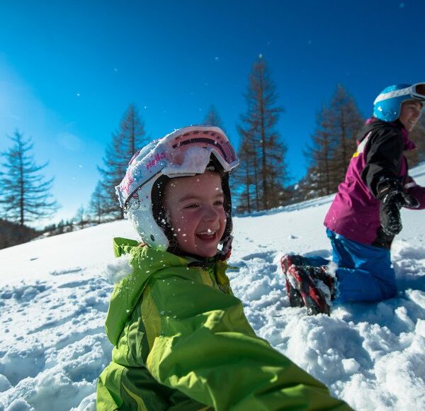 Zwei Kinder mit Helmen und Winterkleidung spielen an einem sonnigen Tag im Schnee, umgeben von Bäumen und Bergen.