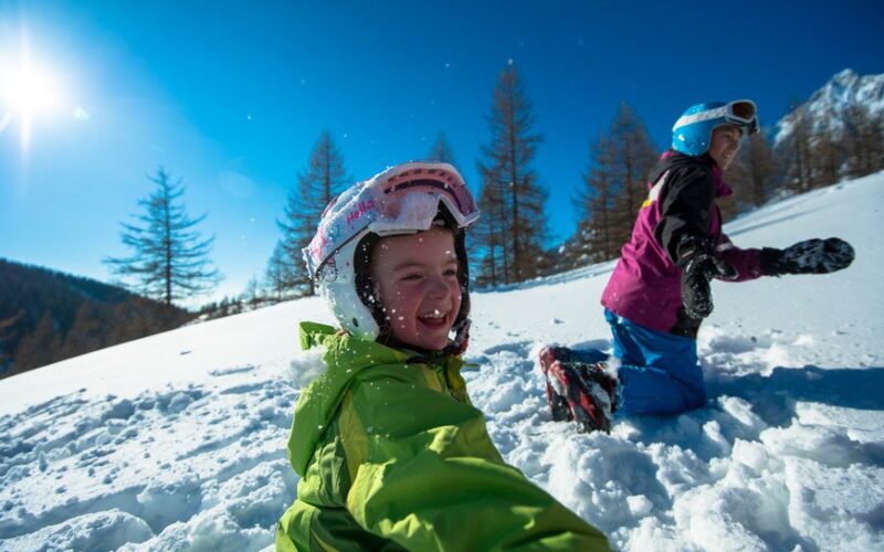 Zwei Kinder mit Helmen und Winterkleidung spielen an einem sonnigen Tag im Schnee, umgeben von Bäumen und Bergen.