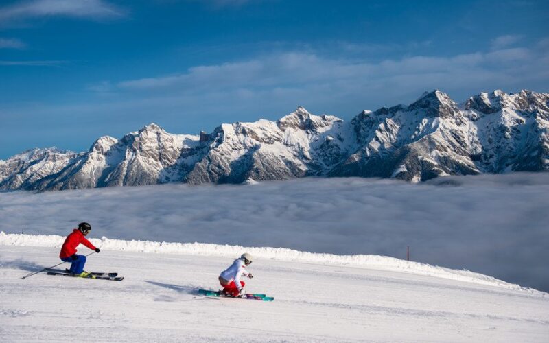 Zwei Skifahrer fahren einen schneebedeckten Hang hinunter, im Hintergrund sind unter einem klaren blauen Himmel eine Bergkette und Wolken zu sehen.