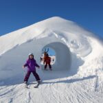 Zwei Kinder in Winterkleidung stehen in der Nähe eines Schneetunnels, in dem sich ein strahlend blauer Himmel und Skispuren befinden.