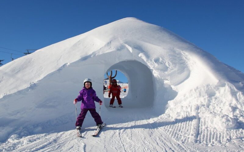 Zwei Kinder in Winterkleidung stehen in der Nähe eines Schneetunnels, in dem sich ein strahlend blauer Himmel und Skispuren befinden.