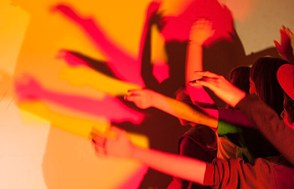 Mehrere Personen heben ihre Arme vor einer Lichtquelle und werfen überlappende, bunte Schatten auf eine Wand.