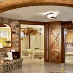 Spa-Interieur mit Steinwänden, Holztüren, gläsernem Dampfbad, Sitzecke, Blumen und einem Tablett mit einem Krug und einem Glas auf einem Tisch. Die warme Beleuchtung schafft eine entspannende Atmosphäre.