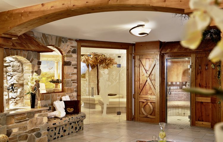 Spa-Interieur mit Steinwänden, Holztüren, gläsernem Dampfbad, Sitzecke, Blumen und einem Tablett mit einem Krug und einem Glas auf einem Tisch. Die warme Beleuchtung schafft eine entspannende Atmosphäre.