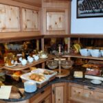 Ein Frühstücksbuffet mit Brötchen, Aufschnitt, Käse, Obst, Joghurt, Bechern und Gläsern, die auf Holzregalen und -theken angeordnet sind.