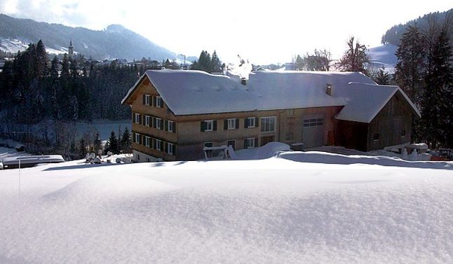 Ein großes Holzgebäude, aus dessen Schornstein Rauch aufsteigt, ist umgeben von tiefem Schnee und Bäumen in einer bergigen Winterlandschaft.