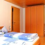 Ein Schlafzimmer mit einem Holzbett, blauem Bettzeug, einem hölzernen Kleiderschrank, einer geschlossenen Tür und einem Holzstuhl an der Wand.