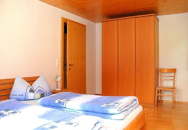 Ein Schlafzimmer mit einem Holzbett, blauem Bettzeug, einem hölzernen Kleiderschrank, einer geschlossenen Tür und einem Holzstuhl an der Wand.
