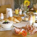 Ein gedeckter Frühstückstisch mit Croissants, gekochten Eiern, Aufschnitt, Käse, Joghurt, Obstsalat, Orangensaft, Müsli, Kaffee und Brot in Körben, mit Blumen als Dekoration.