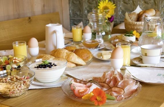 Ein gedeckter Frühstückstisch mit Croissants, gekochten Eiern, Aufschnitt, Käse, Joghurt, Obstsalat, Orangensaft, Müsli, Kaffee und Brot in Körben, mit Blumen als Dekoration.