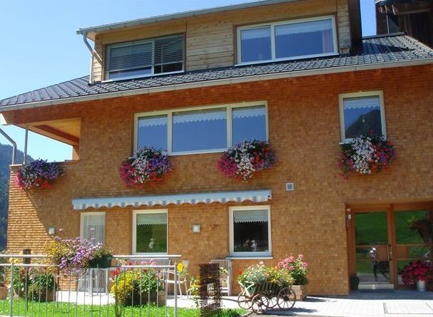 Ein dreistöckiges Haus mit Backsteinfassade und Obergeschoss aus Holz, mit Blumenkästen unter den Fenstern und Topfpflanzen auf der Terrasse.
