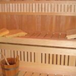 Hölzernes Interieur einer Sauna mit Bänken, einem Eimer und einer Schöpfkelle sowie gefalteten Handtüchern auf der oberen Bank.