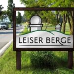 Auf einem Straßenschild steht „Leiser Berge“ und die Grafik zeigt zwei Hügel und zwei Observatoriumskuppeln. Im Hintergrund sind Bäume und ein Lieferwagen zu sehen.