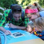 Zwei Erwachsene und zwei Kinder untersuchen an einem sonnigen Tag im Freien, umgeben von Grün und einem Holzzaun, eine Informationstafel genau.