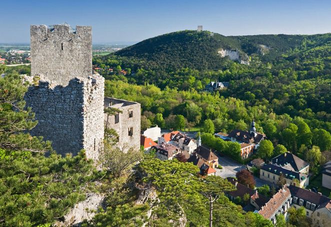 Die Steinruinen einer Burg auf einem Hügel überblicken ein kleines Dorf mit bunten Häusern, umgeben von dichten grünen Wäldern und fernen Hügeln unter einem klaren Himmel.