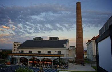 Ein großes Industriegebäude mit einem hohen gemauerten Schornstein, einem Sitzbereich im Freien mit Menschen und einem bewölkten Himmel in der Abenddämmerung.