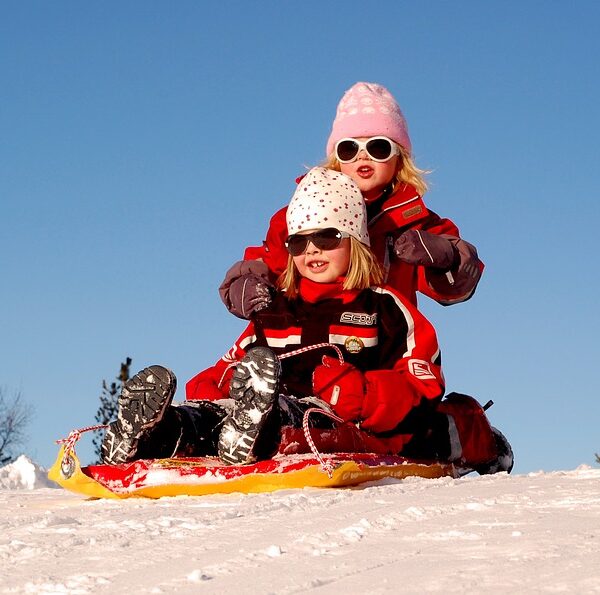 Zwei Kinder in Winterkleidung und mit Sonnenbrille fahren gemeinsam auf einem Schlitten einen verschneiten Hügel hinunter, unter einem klaren blauen Himmel.