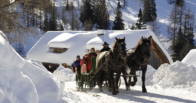 Ein Pferdeschlitten mit mehreren Personen fährt auf einem verschneiten Weg mit schneebedeckten Bäumen und einer Hütte im Hintergrund.