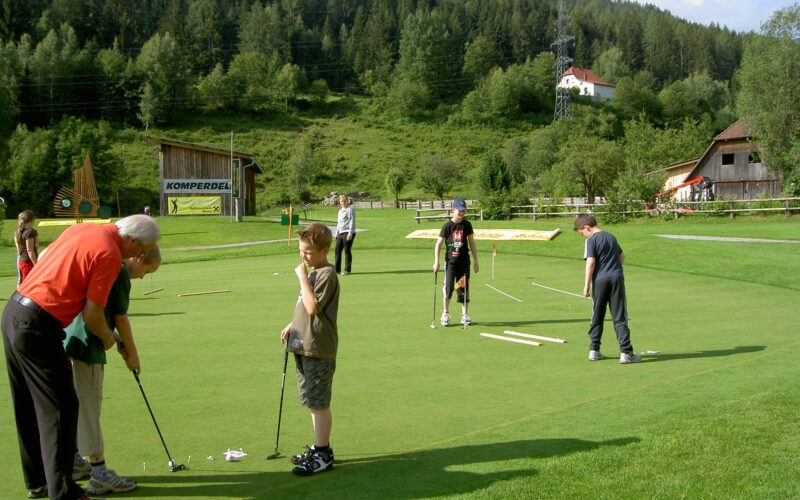 Eine Gruppe von Menschen, darunter Kinder und Erwachsene, übt das Putten auf einem von Bäumen und Gebäuden umgebenen Golfgrün in einer ländlichen Gegend.