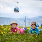 Zwei Kinder liegen auf dem Bauch in einem Feld mit gelben Wildblumen und lächeln in die Kamera, im Hintergrund sind Berge und eine Seilbahn zu sehen.