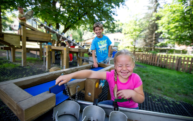 Zwei Kinder spielen an einer Wasseraktivitätsstation auf einem Spielplatz im Freien, im Hintergrund sind Bäume und Holzgeräte zu sehen.