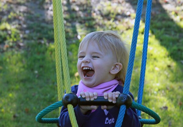 Ein kleines Kind mit blondem Haar lacht, während es sich im Freien an den Seilen einer Schaukel festhält.