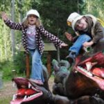 Zwei Kinder mit Hüten posieren spielerisch auf einer großen Drachenskulptur in einer bewaldeten Umgebung im Freien.