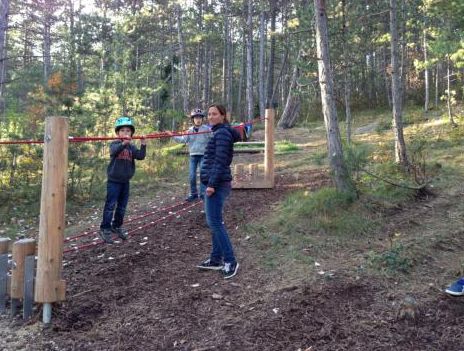 Zwei Kinder mit Helmen bewältigen einen Seilhindernisparcours in einem Waldstück, während ein Erwachsener in der Nähe steht und zusieht.