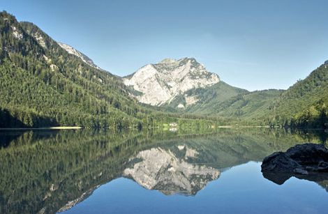 Ein klarer See spiegelt einen bewaldeten Berg unter einem blauen Himmel wider, mit felsigen Hängen und Bäumen entlang der Uferlinie.