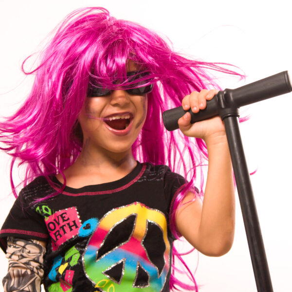 Ein Kind mit rosa Perücke und Sonnenbrille singt in ein Mikrofon, während es ein buntes T-Shirt mit Friedens- und Herzsymbolen trägt.