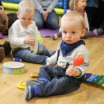 Zwei Kleinkinder sitzen auf dem Boden und spielen mit Musikinstrumenten; eines hält eine Maraca und sitzt neben einem Xylophon, während das andere eine Trommel spielt. Ein großer ausgestopfter Bär ist in der Nähe.