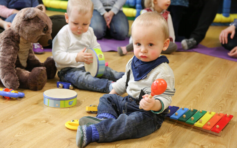 Zwei Kleinkinder sitzen auf dem Boden und spielen mit Musikinstrumenten; eines hält eine Maraca und sitzt neben einem Xylophon, während das andere eine Trommel spielt. Ein großer ausgestopfter Bär ist in der Nähe.