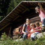 Eine vierköpfige Familie sitzt auf einem Baumstamm vor einer Holzhütte im Wald; der Vater zeigt auf etwas in der Ferne.