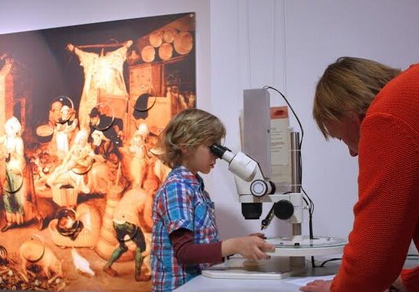 Ein Kind schaut in ein Mikroskop, während ein Erwachsener zusieht. Im Hintergrund ist an der Wand ein buntes Gemälde zu sehen.