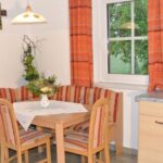 Eine kleine Küchenecke mit einem Holztisch, einer gestreiften Polsterbank und Stühlen, orangefarbenen Vorhängen, einem Fenster, einer Wanddekoration in Form eines Kreuzes und einer einfachen Arbeitsplatte mit einem Waschbecken.