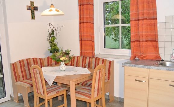 Eine kleine Küchenecke mit einem Holztisch, einer gestreiften Polsterbank und Stühlen, orangefarbenen Vorhängen, einem Fenster, einer Wanddekoration in Form eines Kreuzes und einer einfachen Arbeitsplatte mit einem Waschbecken.