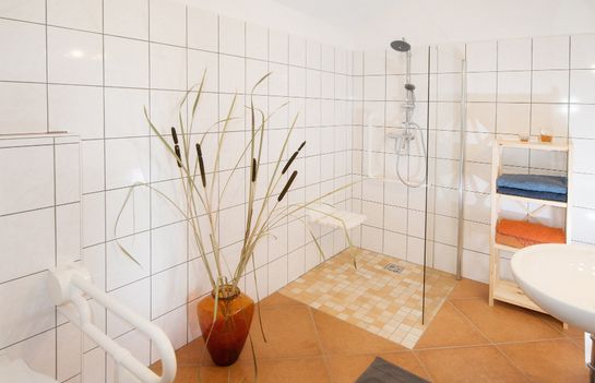 Ein gefliestes Badezimmer mit einer begehbaren Dusche, einer Glaswand, einem an der Wand befestigten Duschsitz, einer Topfpflanze, einem Regal mit gefalteten Handtüchern und einem weißen Waschbecken.