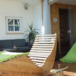 Ein sonnendurchfluteter Aufenthaltsraum mit einem hölzernen Lehnstuhl, grünen Sitzsäcken, einem Sofa, Topfpflanzen und einem kleinen Saunaraum im Hintergrund.