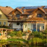 Ein zweistöckiges Haus mit Sonnenkollektoren und einem begrünten Dach steht an einem sonnigen Tag neben einem Teich und einer Spielplatzrutsche, direkt neben einem gelben Gebäude.