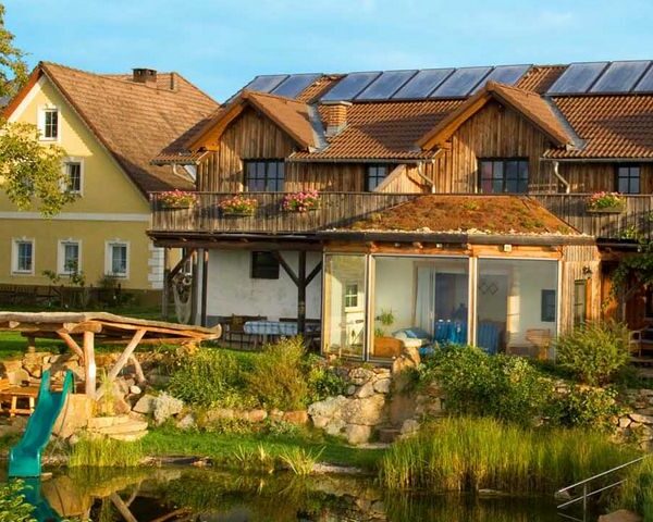 Ein zweistöckiges Haus mit Sonnenkollektoren und einem begrünten Dach steht an einem sonnigen Tag neben einem Teich und einer Spielplatzrutsche, direkt neben einem gelben Gebäude.