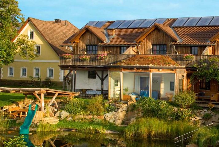 Ein zweistöckiges Haus mit Sonnenkollektoren und einem begrünten Dach steht an einem sonnigen Tag neben einem Teich und einer Spielplatzrutsche, direkt neben einem gelben Gebäude.